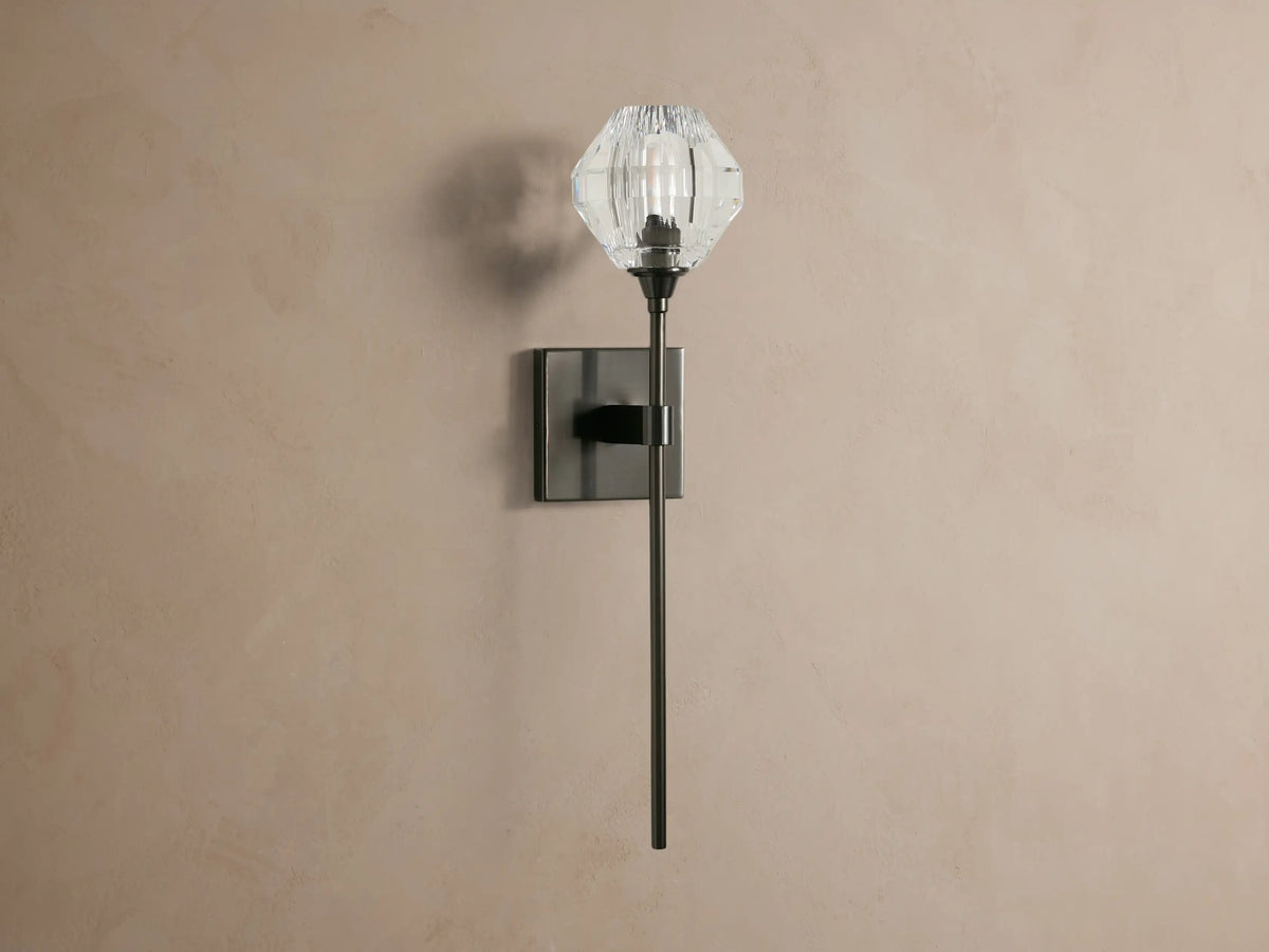 Amoroso Sconce with a clear faceted crystal shade & lit bulb. Features a dark metal arm & square backplate on a tan wall.