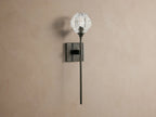 Amoroso Sconce with a clear faceted crystal shade & lit bulb. Features a dark metal arm & square backplate on a tan wall.