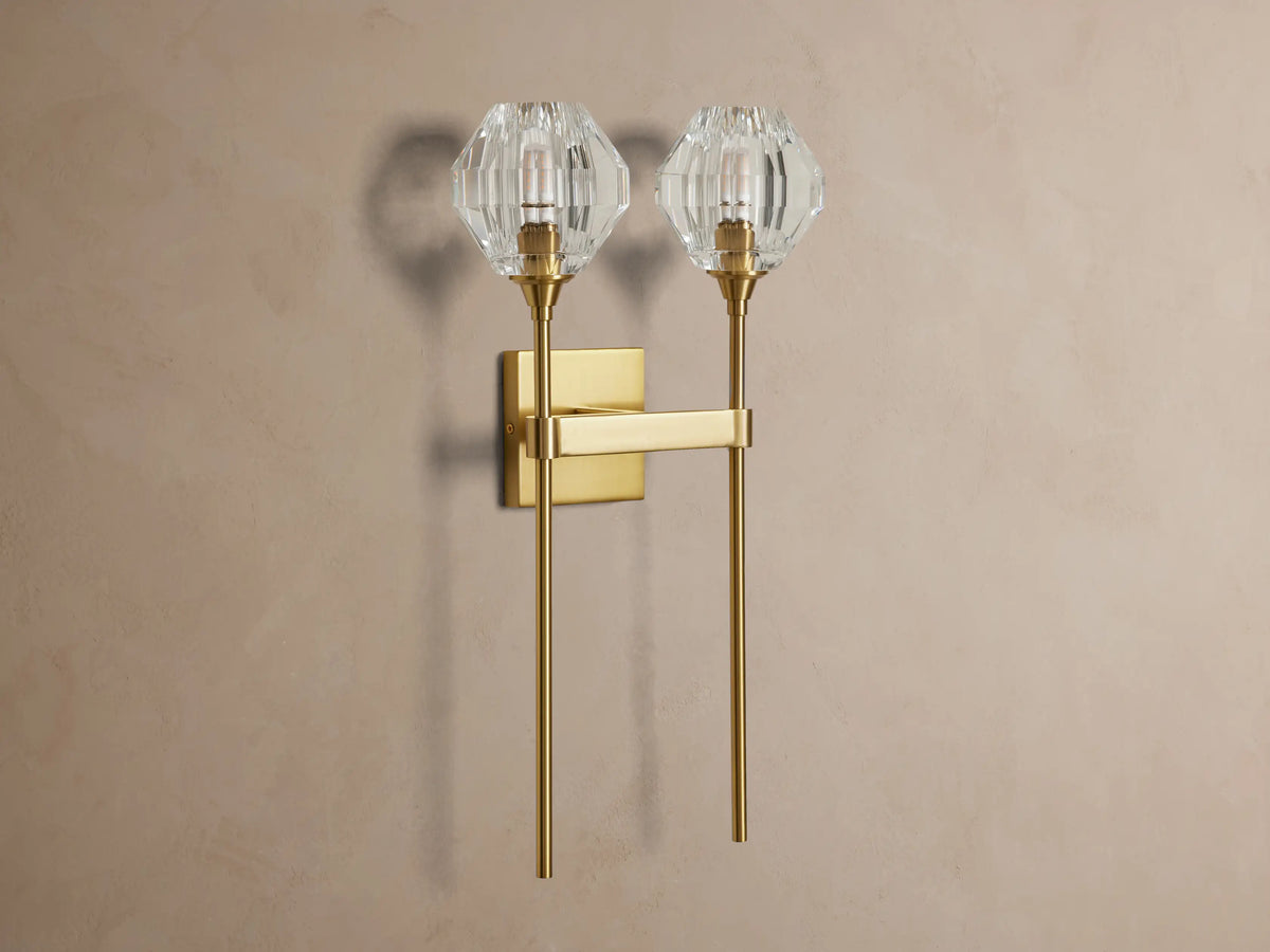 Amoroso Double Sconce, brass-plated with two illuminated faceted crystal shades, vertically mounted on a taupe wall.