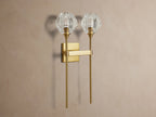 Amoroso Double Sconce, brass-plated with two illuminated faceted crystal shades, vertically mounted on a taupe wall.