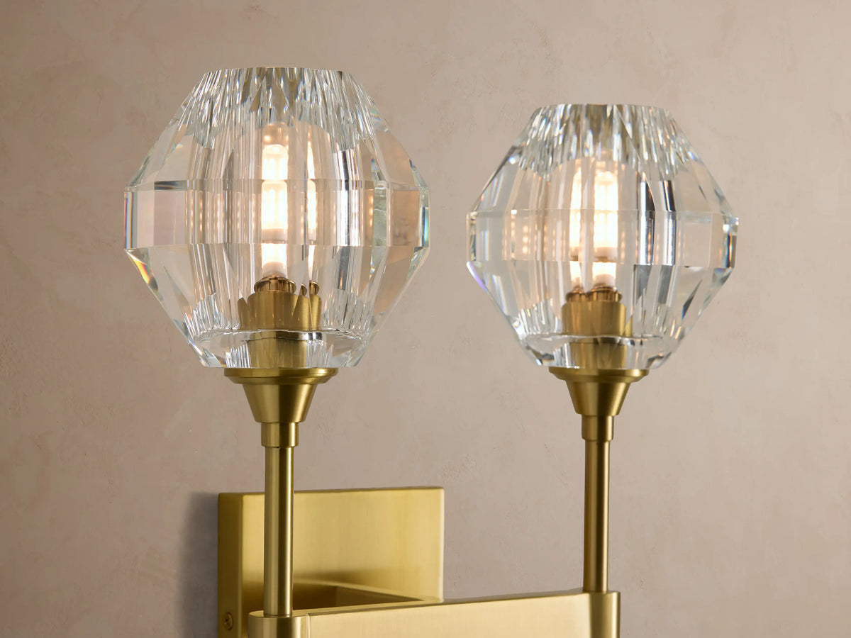 Amoroso Double Sconce with two faceted crystal shades & brass-plated arms, mounted vertically on a light beige wall.