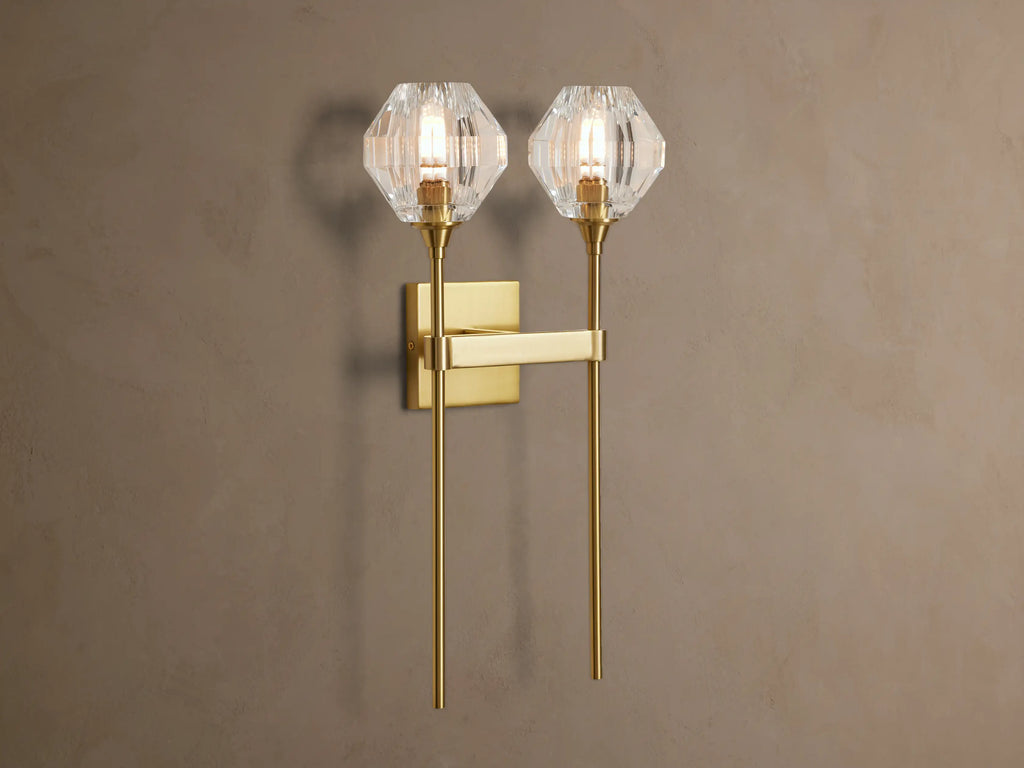 Amoroso Double Sconce, brass-plated with two illuminated faceted crystal shades, vertically mounted on a taupe wall.
