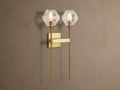 Amoroso Double Sconce, brass-plated with two illuminated faceted crystal shades, vertically mounted on a taupe wall.