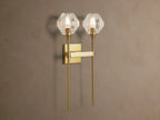 Amoroso Double Sconce, brass-plated with two illuminated faceted crystal shades, vertically mounted on a taupe wall.