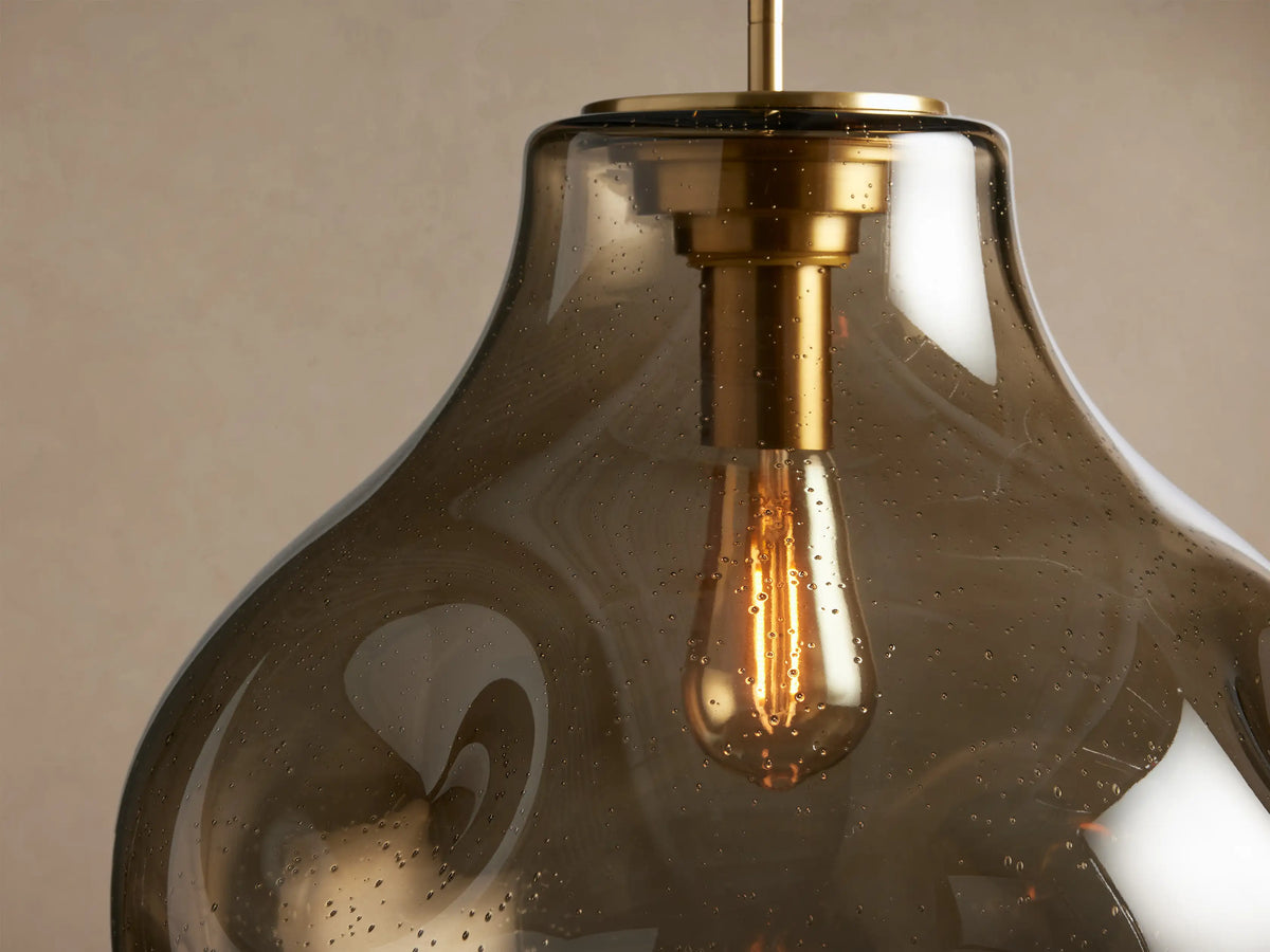 Organic 16" Pendant: Smoky gray, organic mouth-blown glass, brass hardware, & exposed Edison bulb hanging on a neutral wall.