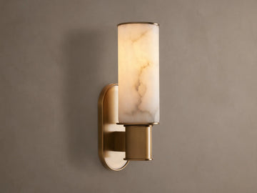 Harlin Sconce in Alabaster with a cylindrical cloche & burnished brass base, glowing warmly against a gray wall.