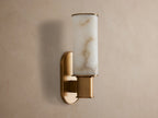 Harlin Sconce in Alabaster with a cylindrical cloche & burnished brass base, glowing warmly against a gray wall.