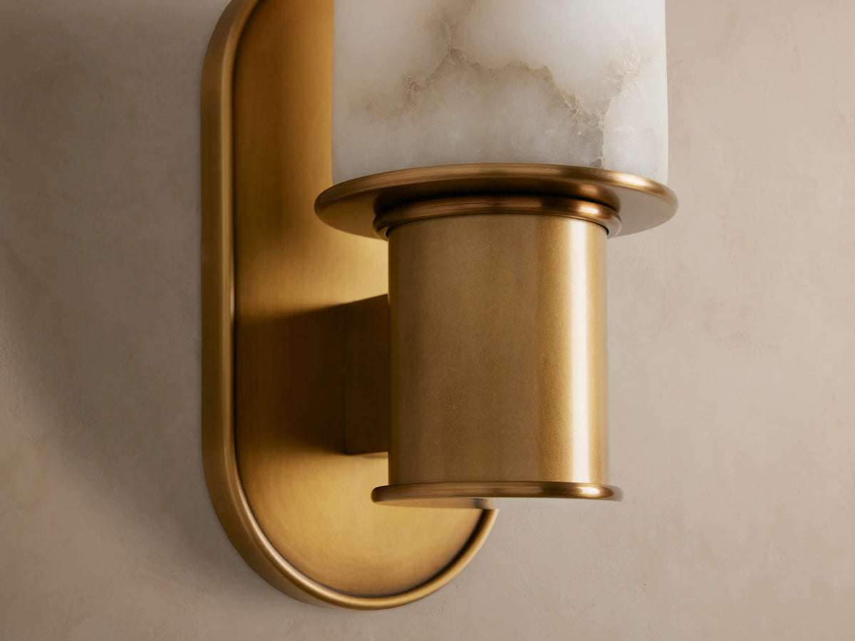 Vertical Harlin Sconce with cylindrical white alabaster shade, burnished brass base & racetrack backplate on a beige wall.