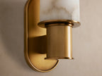 Vertical Harlin Sconce with cylindrical white alabaster shade, burnished brass base & racetrack backplate on a beige wall.