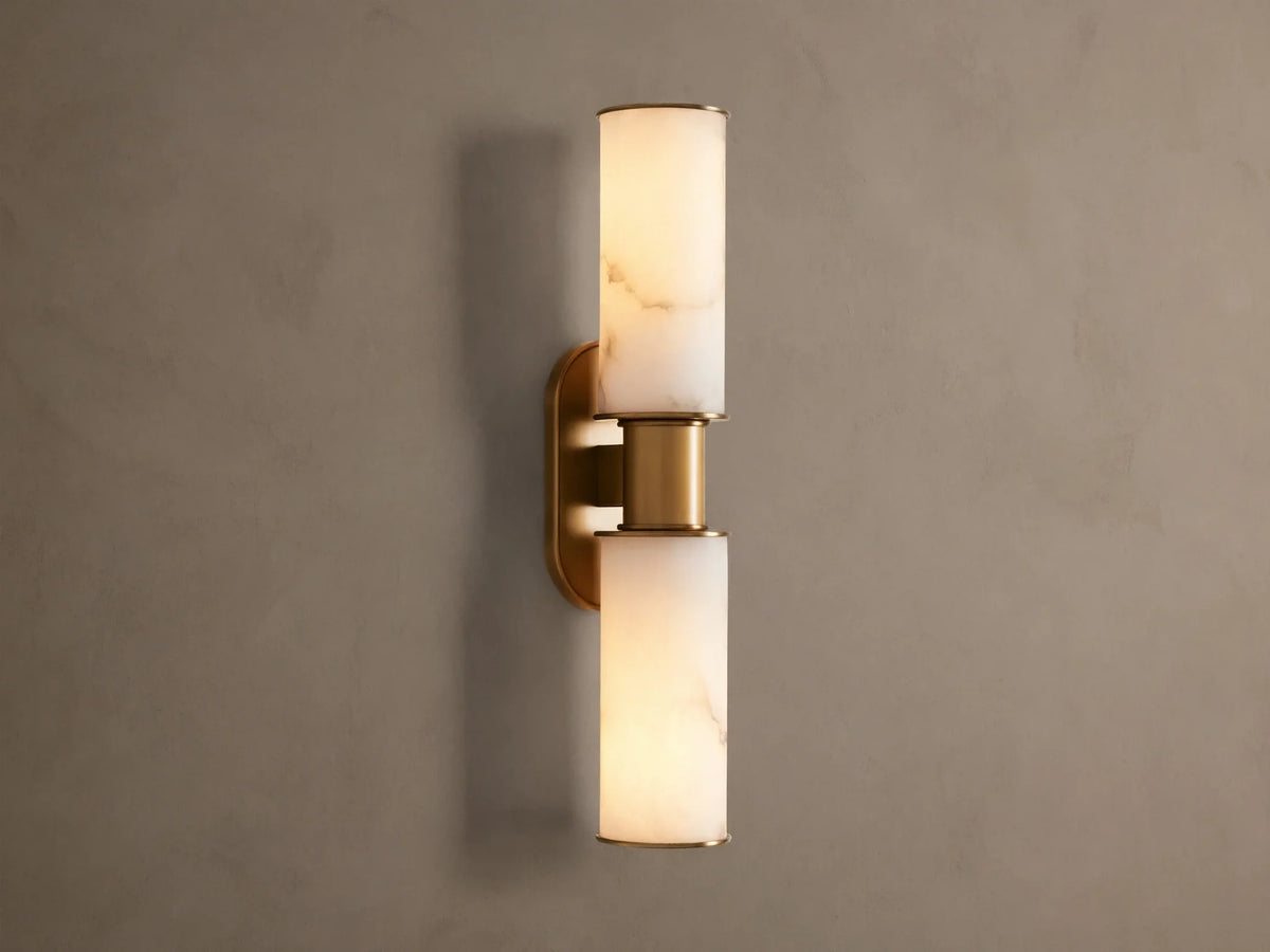 Harlin Double Sconce: Two glowing alabaster cylinder shades with burnished brass trim, mounted vertically on a neutral wall.
