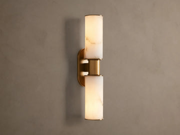 Harlin Double Sconce: Two glowing alabaster cylinder shades with burnished brass trim, mounted vertically on a neutral wall.