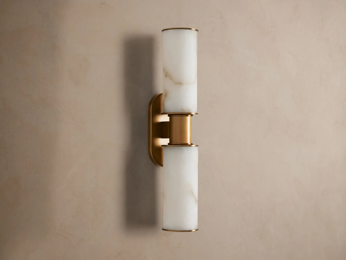 Harlin Double Sconce: Two glowing alabaster cylinder shades with burnished brass trim, mounted vertically on a neutral wall.