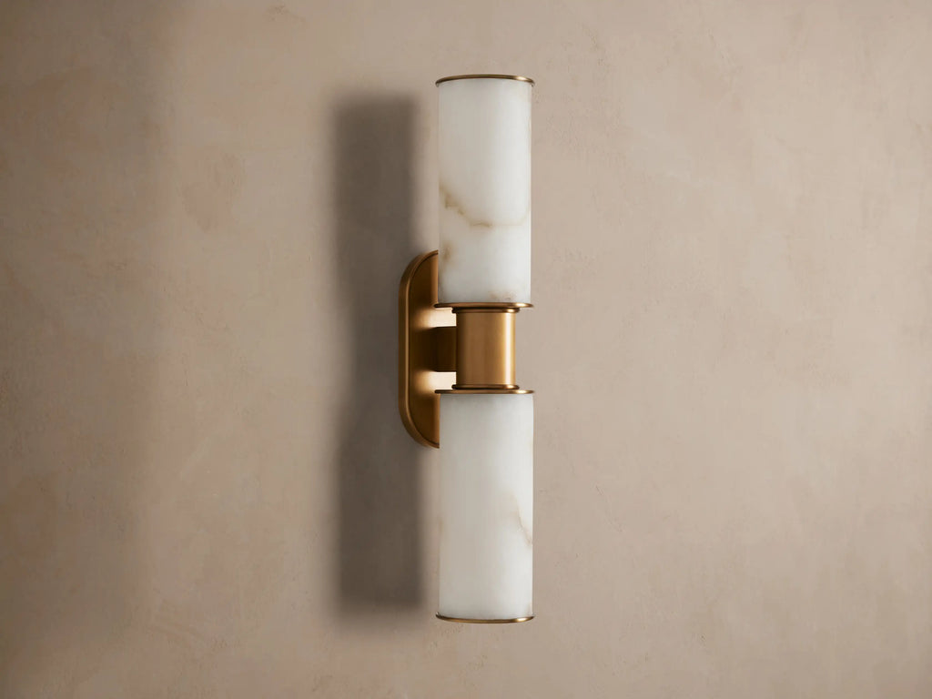 Harlin Double Sconce: Two glowing alabaster cylinder shades with burnished brass trim, mounted vertically on a neutral wall.