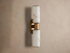 Harlin Double Sconce: Two glowing alabaster cylinder shades with burnished brass trim, mounted vertically on a neutral wall.