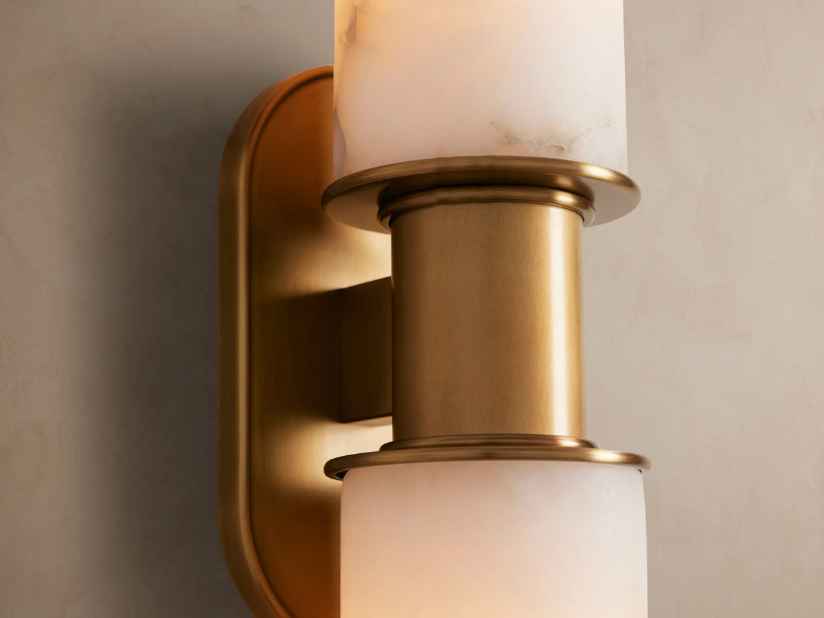 Harlin Double Sconce featuring two vertical white alabaster cloches and burnished brass accents on a light beige wall.
