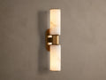 Harlin Double Sconce: Two glowing alabaster cylinder shades with burnished brass trim, mounted vertically on a neutral wall.