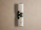 Harlin Double Sconce in Alabaster. Bronze plated fixture with two glowing alabaster cylindrical shades on a taupe wall.