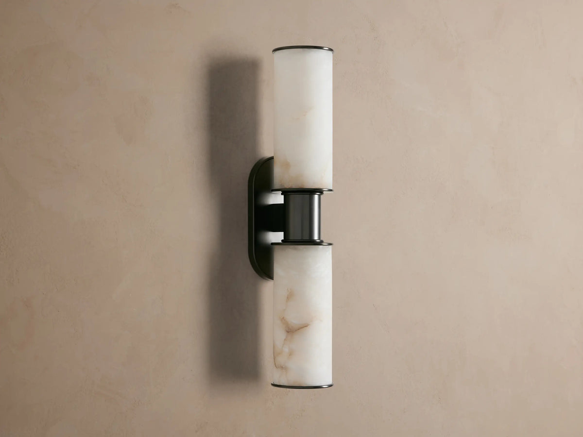 Harlin Double Sconce in Alabaster. Bronze plated fixture with two glowing alabaster cylindrical shades on a taupe wall.