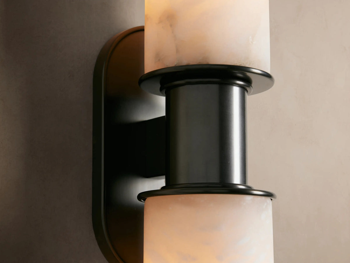 Harlin Double Sconce featuring two stacked alabaster cloches with natural veining on a vertical dark bronze backplate, on a tan wall.