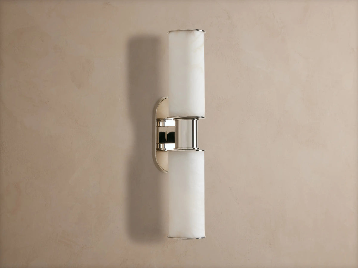 Vertical Harlin Double Sconce in Alabaster with two glowing cylindrical shades and polished nickel on a taupe wall.
