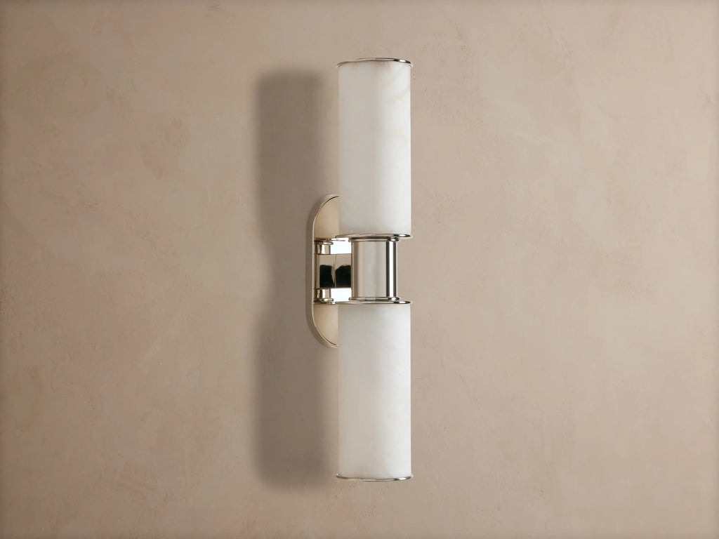 Vertical Harlin Double Sconce in Alabaster with two glowing cylindrical shades and polished nickel on a taupe wall.
