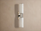Vertical Harlin Double Sconce in Alabaster with two glowing cylindrical shades and polished nickel on a taupe wall.