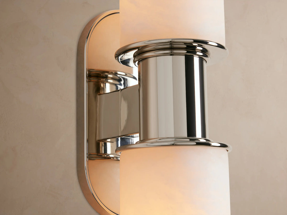 Harlin Double Sconce with two cylindrical white alabaster shades, polished nickel central support & oval backplate, on a beige wall.