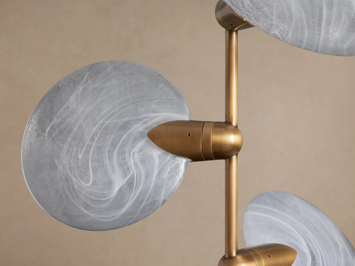 Lyla Pendant: Vertical brass rod with seven translucent white, swirled hand-blown glass discs angled outwards, against a beige wall.