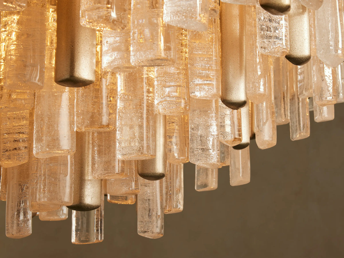 Neve Round Chandelier close-up showing textured clear & bronze cast glass pieces and champagne brass metallic rods hanging. Warm light.
