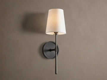 Rhodes Sconce with tapered linen shade on a dark bronze arm and circular backplate, mounted on a light brown wall.
