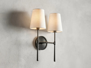 Rhodes Double Sconce with bronze finish, dual tapered linen shades, mounted on a circular backplate, lit on a beige wall.