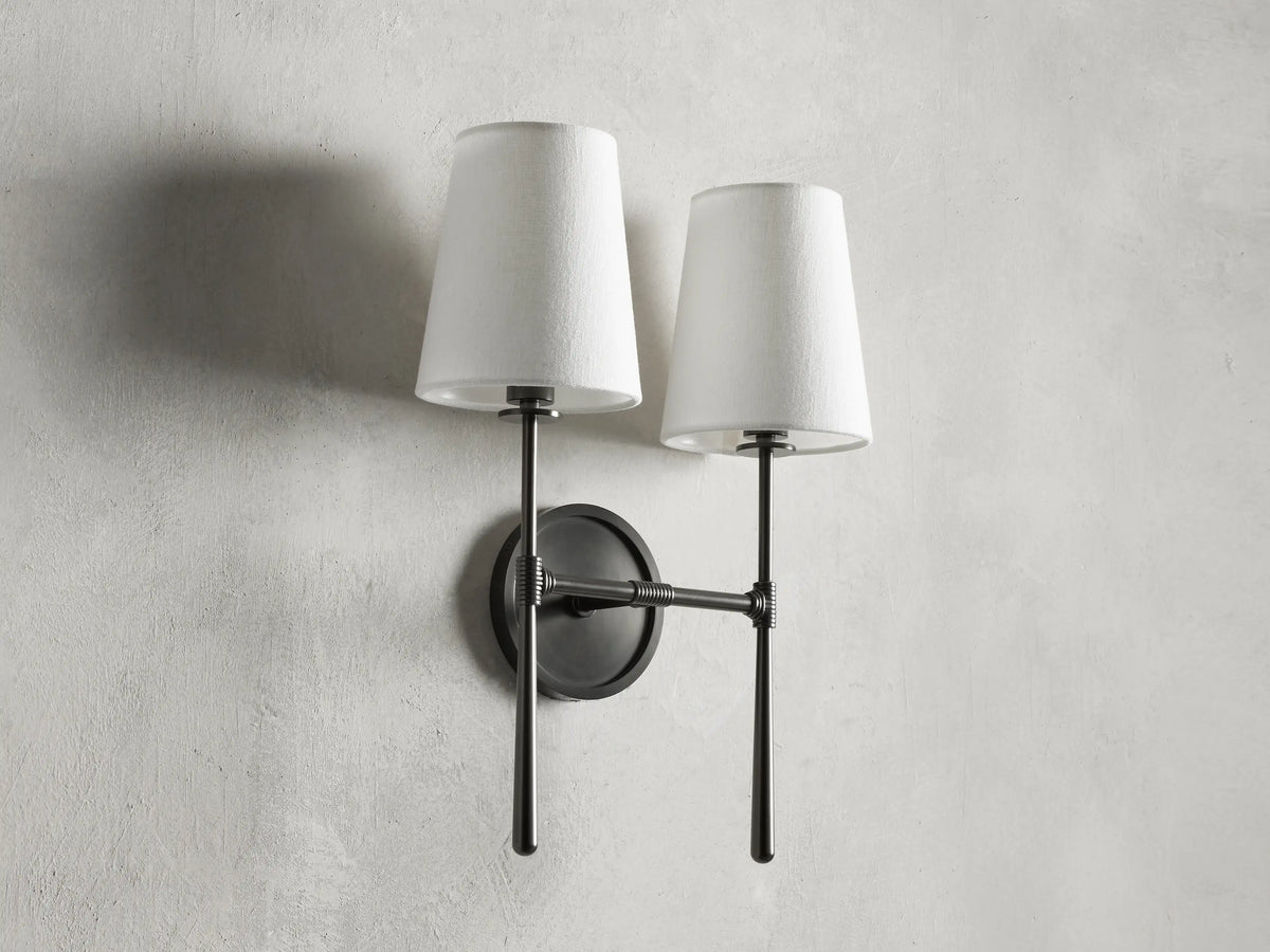 Rhodes Double Sconce with bronze finish, two white tapered linen shades, mounted on a round backplate against a light wall.