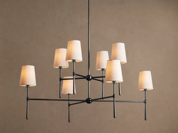 Rhodes Chandelier with 8 light beige tapered linen shades on a dark metal frame, hanging against a tan wall.