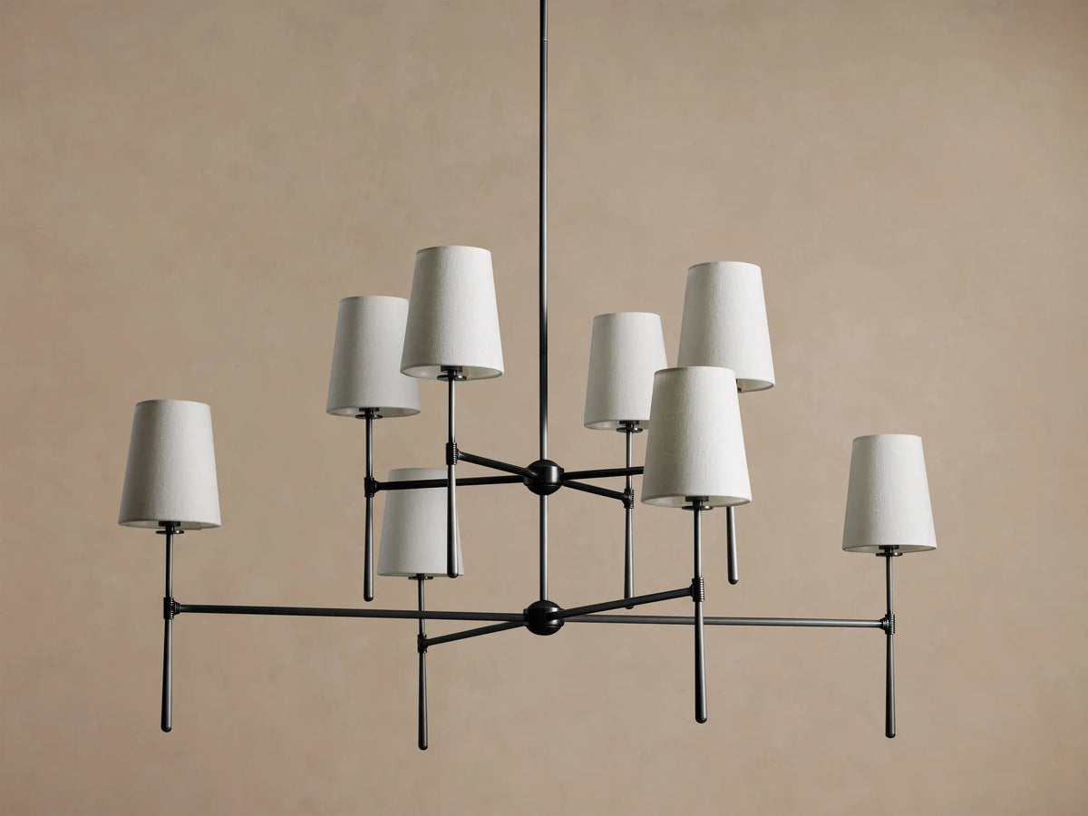 Rhodes Chandelier with 8 light beige tapered linen shades on a dark metal frame, hanging against a tan wall.