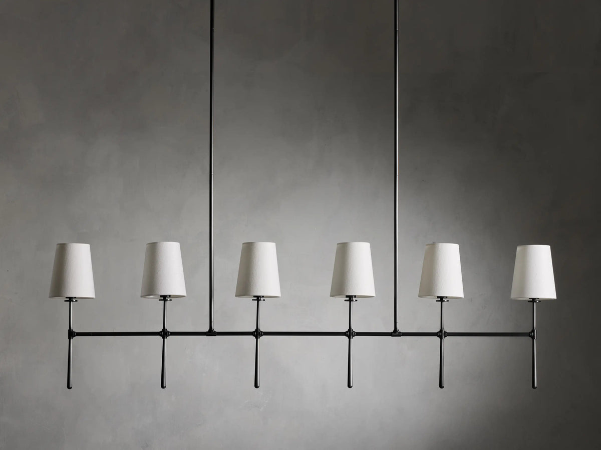 Rhodes Linear Chandelier with six tapered linen shades on a bronze-plated steel frame, suspended against a neutral wall.