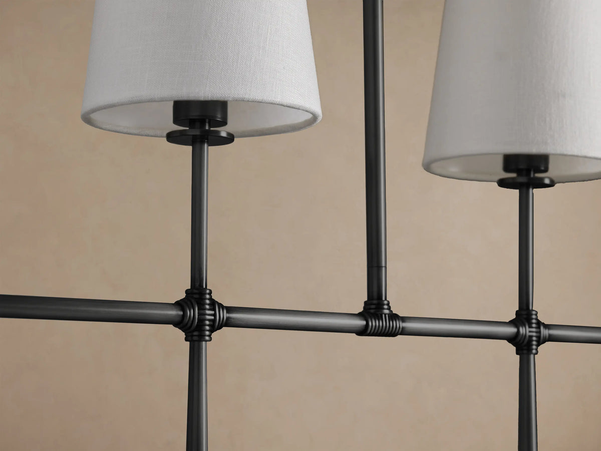 Rhodes Linear Chandelier with 5 tapered off-white linen shades on a dark linear metal frame, suspended by two rods.