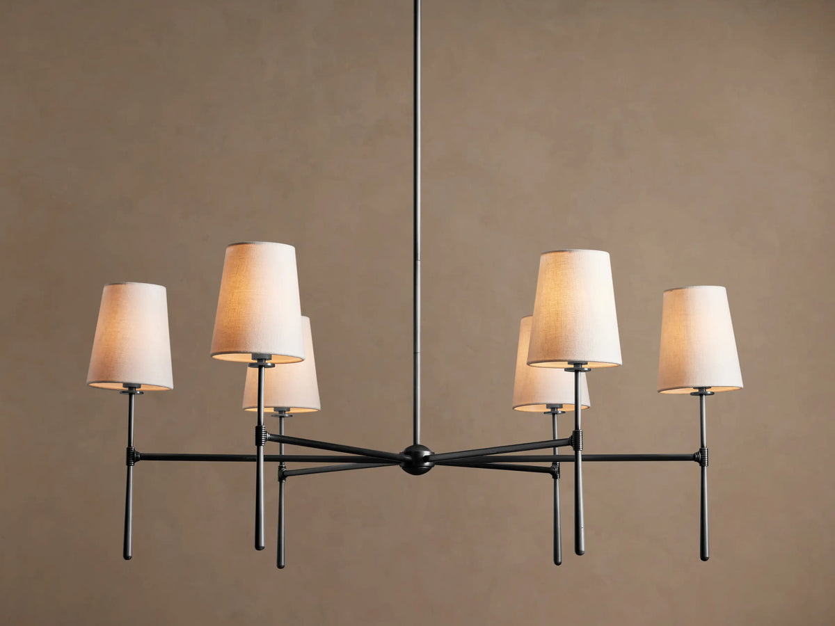 Rhodes Round Chandelier, dark metal frame with six lit tapered linen shades, centered against a warm brown wall.