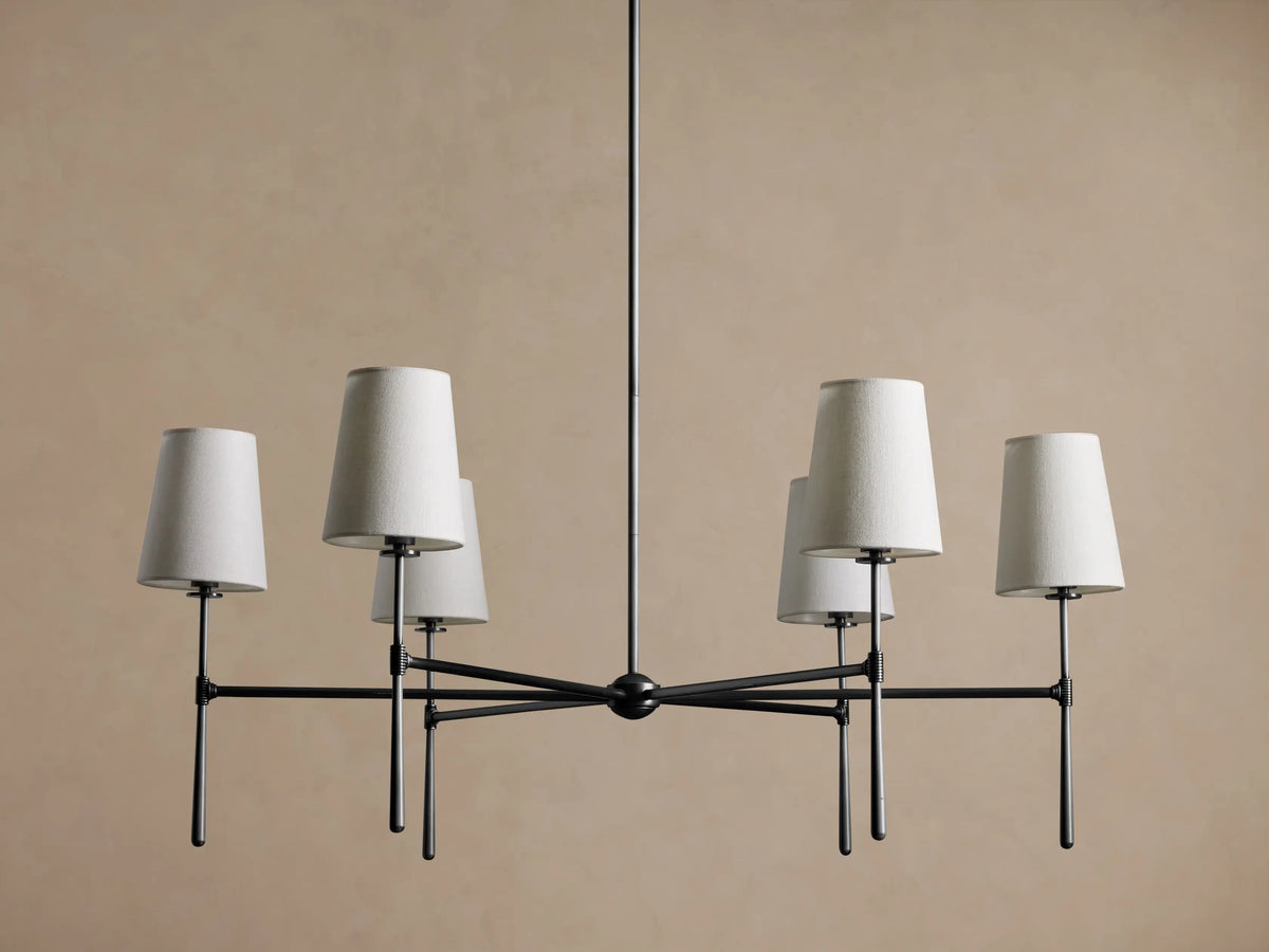 Rhodes Round Chandelier, dark metal frame with six lit tapered linen shades, centered against a warm brown wall.