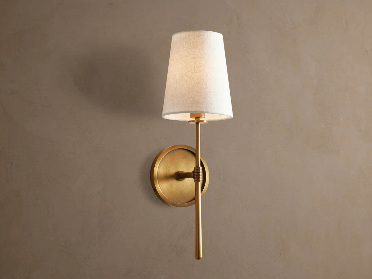 Rhodes Sconce: burnished brass finish wall light, round backplate, and a lit, tapered light linen shade, mounted on a taupe wall.
