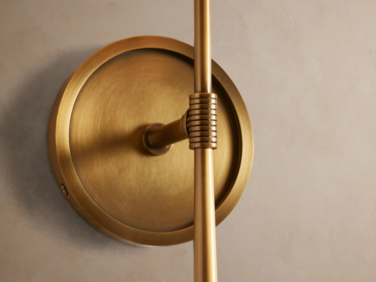Rhodes Sconce in burnished brass, mounted on a light beige wall, with an off-white tapered linen shade.