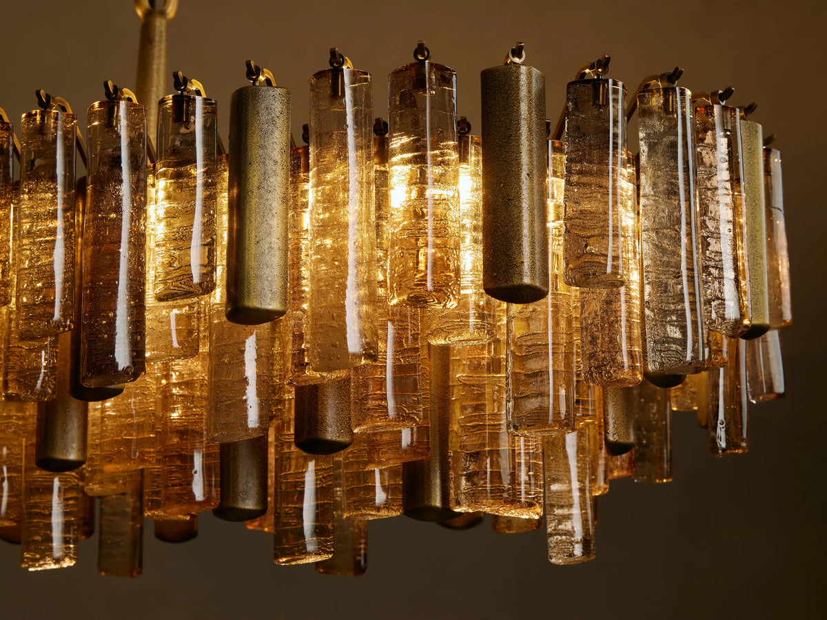 Neve Linear Chandelier close-up: amber textured cast glass and brass cylinders hang vertically, glowing warmly from within.