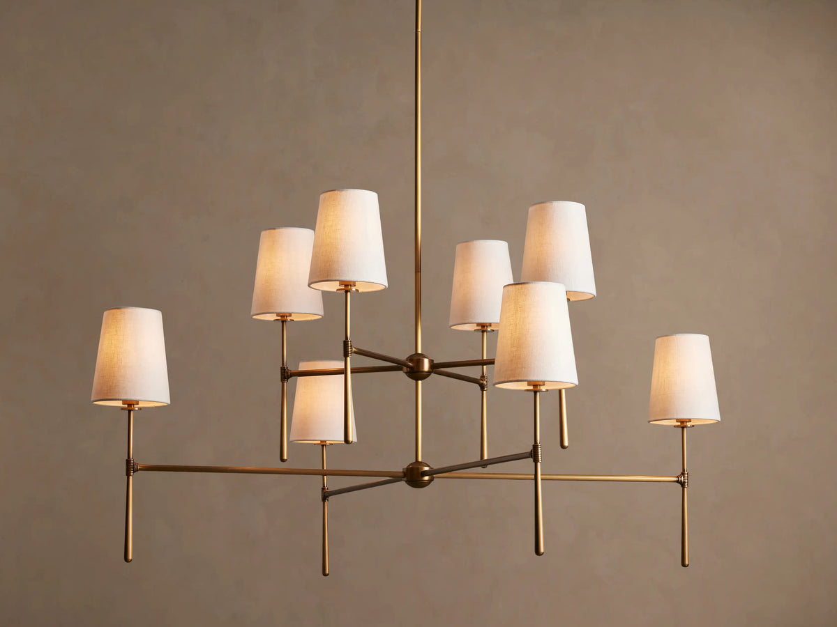Rhodes Chandelier with burnished brass frame, featuring eight tapered off-white linen shades emitting a warm light.
