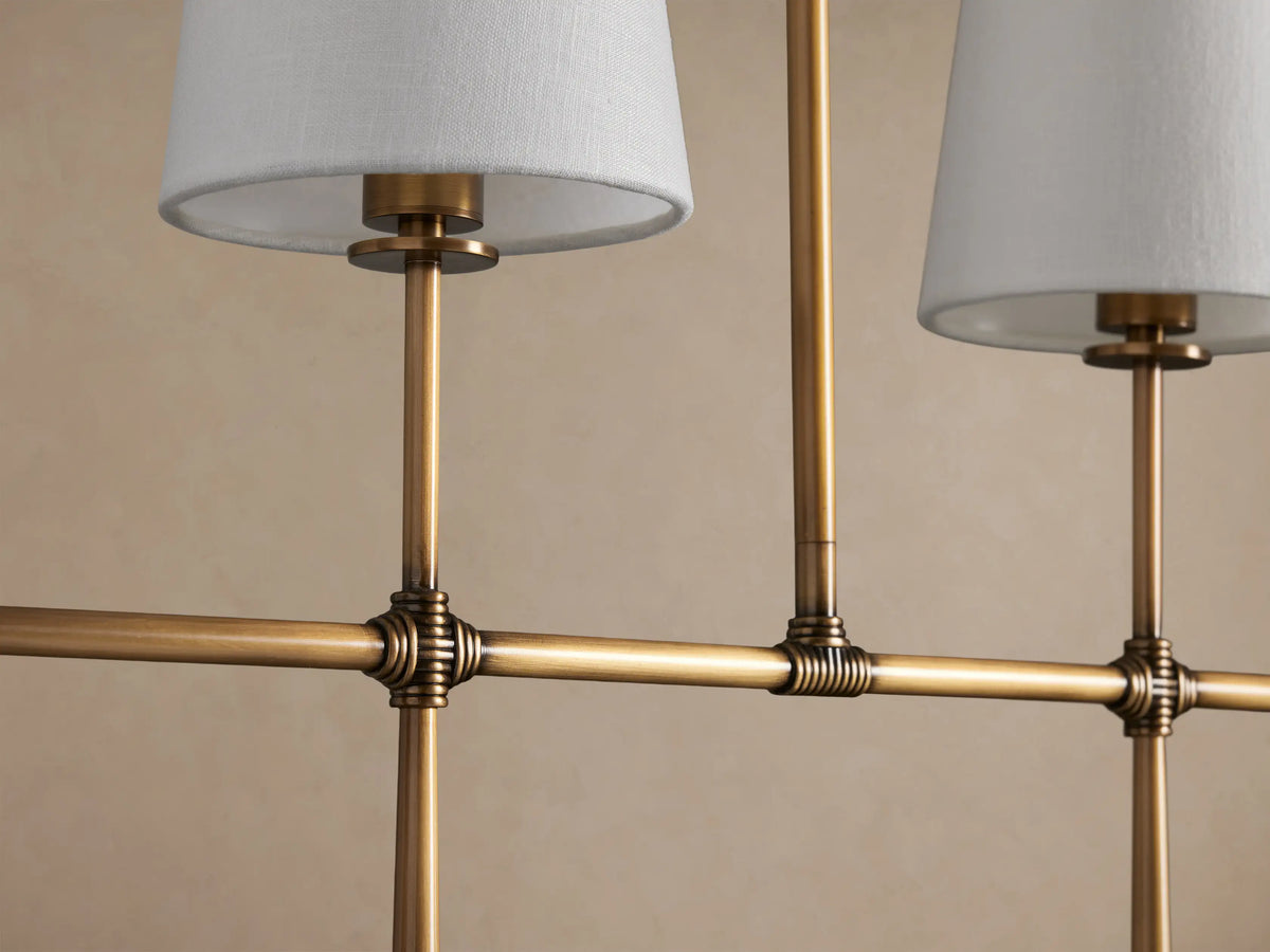 Rhodes Linear Chandelier with 5 white tapered linen shades on a burnished brass linear frame, against a plain light brown background.