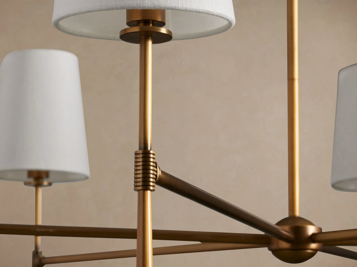 Rhodes Round Chandelier with 6 lights, burnished brass frame, and tapered off-white linen shades, hanging against a light wall.