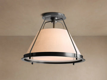 Shelton Flush Mount ceiling light with dark metal frame and large, tapered cream linen shade glowing warmly, mounted on a beige ceiling.