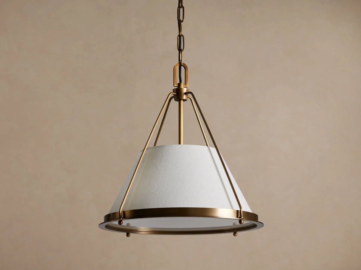 Shelton Pendant: burnished brass metal frame & chain, holds glowing tapered off-white linen shade, hanging against beige wall.