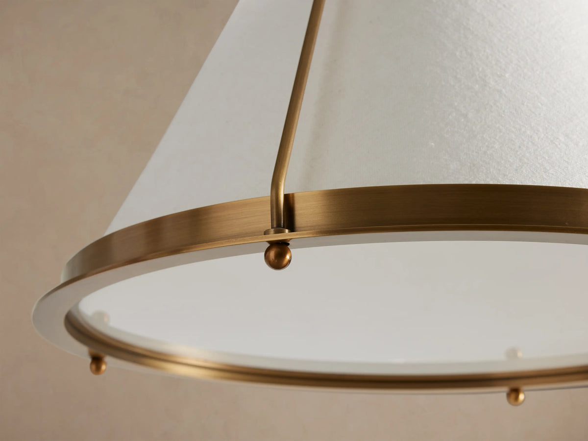 Shelton Pendant light: brass-finished metal frame holding a tapered white linen shade, with a bottom diffuser, hanging against a light brown wall.