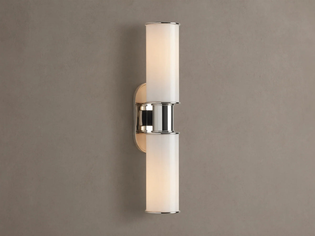 Harlin Double Sconce with two glowing vertical milk glass shades and polished nickel accents, mounted on a textured brown wall.