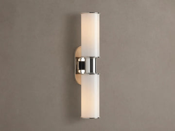 Harlin Double Sconce with two glowing vertical milk glass shades and polished nickel accents, mounted on a textured brown wall.