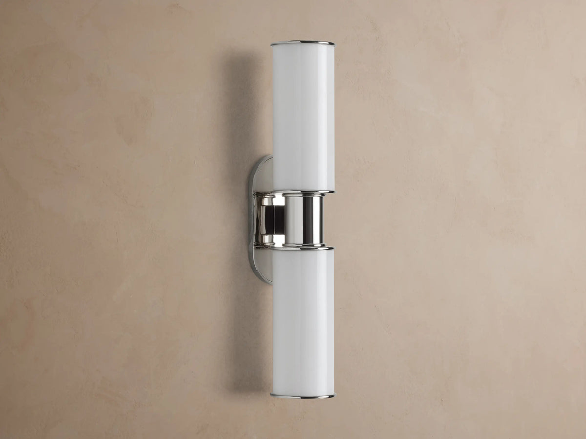 Harlin Double Sconce with two glowing vertical milk glass shades and polished nickel accents, mounted on a textured brown wall.